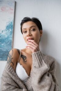 Sensual tattooed artist looking at camera near painting on wall in workshop