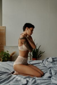 side view of seductive tattooed woman in lingerie embracing herself while sitting on bed