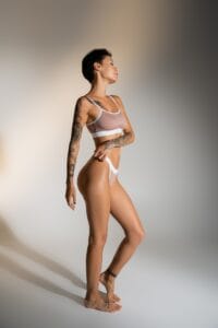 side view of slim brunette woman with tattooed body posing in lingerie on grey background