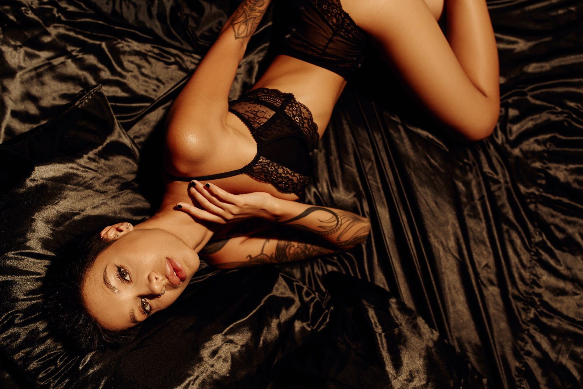 top view of sexy tattooed woman in lace lingerie lying on black silk bedding and looking at camera